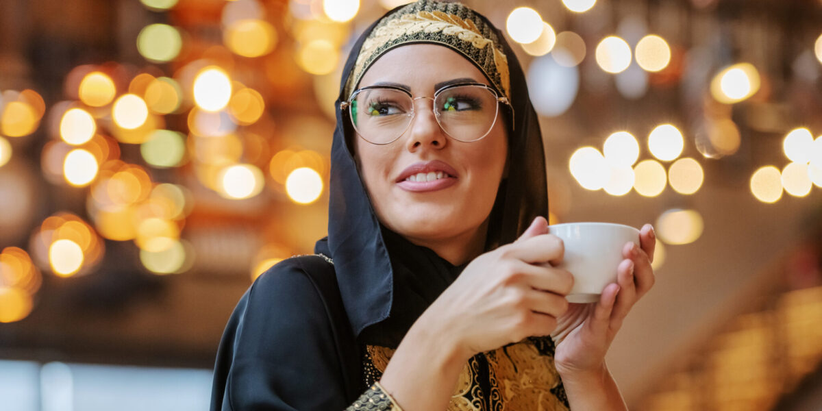 Attractive positive smiling muslim woman in traditional wear sitting in cafe alone, holding cup of fresh coffee and looking away.