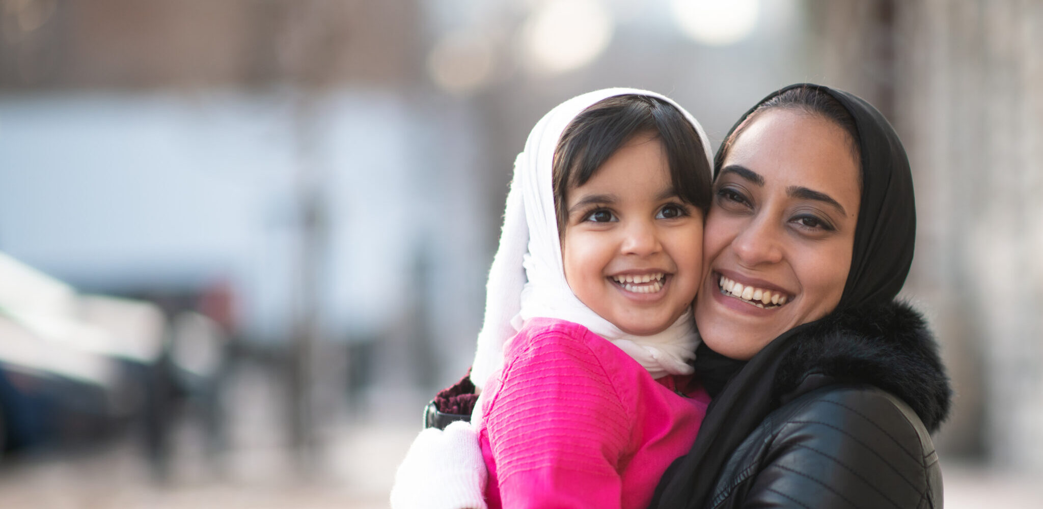 A beautiful young Muslim mother holds her daughter in a loving embrace. They are both smiling and facing forward while outside one day during the winter season.